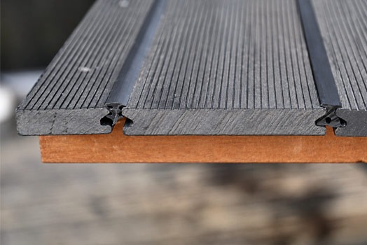 Detailaufnahme nachrüstbarer schwarzer AP TPE-Fugenprofile zwischen gerillten, anthrazitfarbenen WPC-Terrassendielen. Die Nahaufnahme zeigt zwei eingesetzte elastische Dichtprofile mit unterschiedlichen Profilquerschnitten, die die Dielenfugen von oben abdecken. Die natürliche Oberflächenstruktur der Terrassendielen und die darunterliegende Holzunterkonstruktion sind deutlich erkennbar.