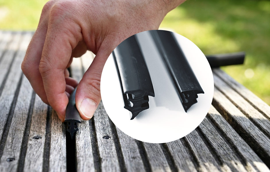 Fugenprofil | Dichtprofil | Nahaufnahme der einfachen Nachrüstung eines schwarzen AP TPE-Fugenprofils in einer gerillten Holzterrasse: Eine Hand drückt ganz unkompliziert das elastische Dichtprofil von oben in die offene Fuge zwischen zwei Terrassendielen. Eine kreisförmige Detailansicht zeigt zwei unterschiedlich große TPE-Fugenprofile mit ihren speziellen Querschnitten. Die nachrüstbaren Kunststoffprofile dienen zum Schutz der Terrassenfugen vor Laub, Tannennadeln und Schmutz.